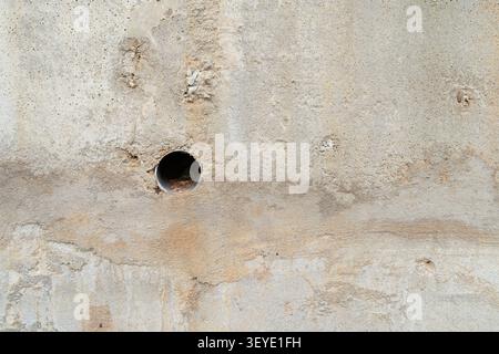 A weathered concrete wall with a small circular drainage hole, featuring rough textures, stains, and natural aging patterns. Stock Photo