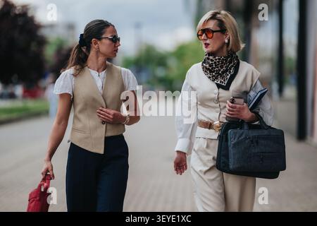 Two businesswomen walking and talking on a city sidewalk in professional attire Stock Photo
