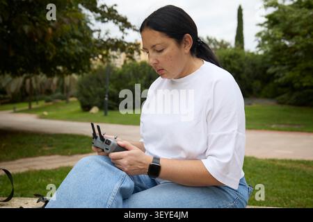 Woman sitting on a stone bench, flying drone using a remote controller in a green city park. Stock Photo