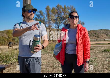 Man and woman walking together along a sunny nature trail with backpacks and casual outdoor clothing. Stock Photo