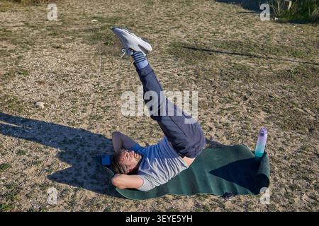 A man performing a vertical leg raise while lying on a fitness mat during an outdoor workout session. Stock Photo