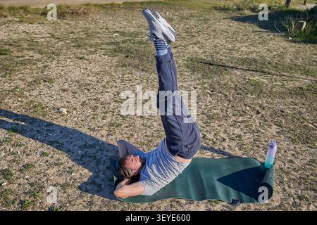 A man performing a vertical leg raise while lying on a fitness mat during an outdoor workout session. Stock Photo