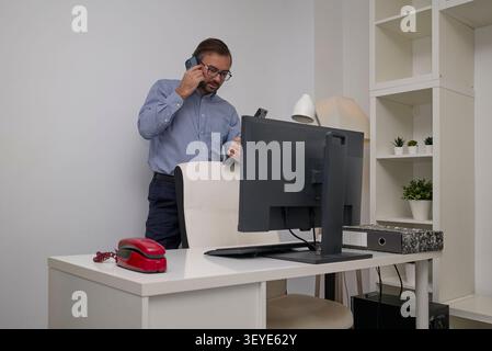 A businessman multitasks in a modern office, speaking on a smartphone while checking documents and working at a desk. Stock Photo