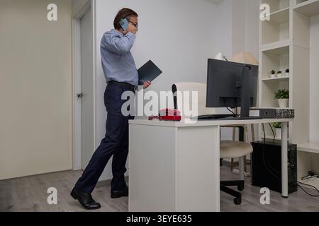 A businessman in formal clothing stands in a modern office, speaking on a smartphone while holding a folder. Stock Photo