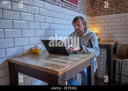 Man sitting at a wooden table in a cozy cafe using a laptop, surrounded by white tiles and warm decorative lighting. Stock Photo