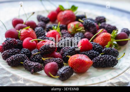 Fresh strawberries, mulberries, and cherries displayed on a decorative plate with green leaves, showcasing a vibrant assortment of berries in a bright Stock Photo