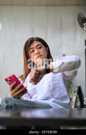 Southeast Asian Thai transgender businesswoman in her 40s sits at a low angle view table while checking smartwatch notifications and using phone. She Stock Photo