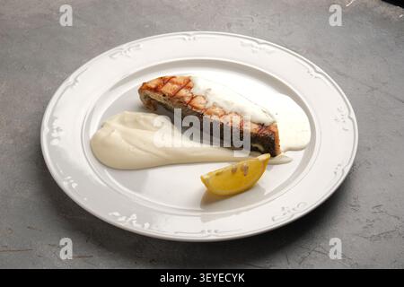 Grilled fish fillet with creamy sauce and lemon wedge on white plate over stone Stock Photo