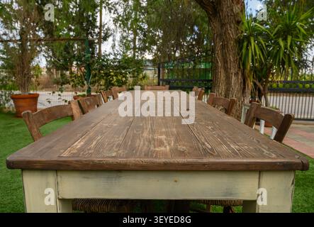 A weathered wooden table lined with rustic chairs sits in a lush garden bathed in warm sunlight. Stock Photo