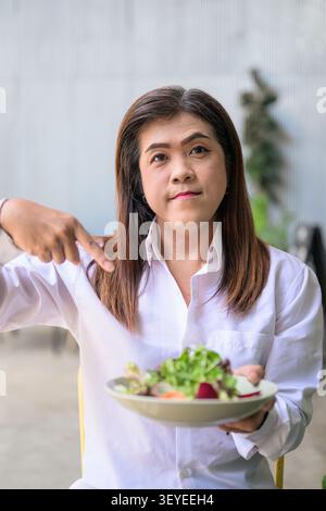 Southeast Asian Thai transgender businesswoman in her 40s sits at an outdoor terrace table while pointing her index finger down at a fresh salad bowl. Stock Photo