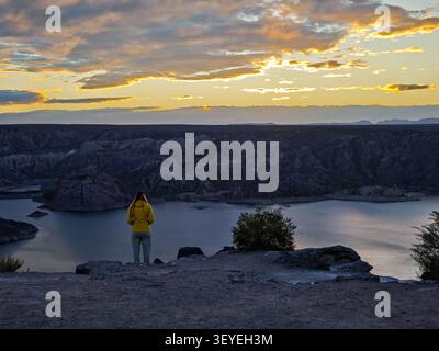 Back view of a female traveler in a yellow jacket overlooking a scenic mountain lake at sunset Stock Photo