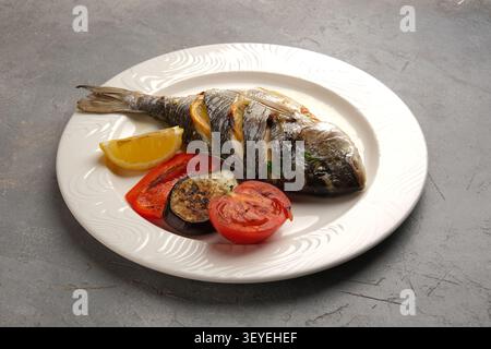 Roasted whole fish served on white plate with grilled vegetables and lemon wedge Stock Photo