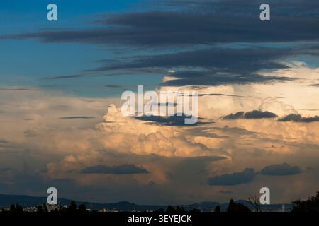Stunning formation of cumulonimbus clouds illuminated by the setting sun in a dramatic sky above a distant city silhouette. Stock Photo