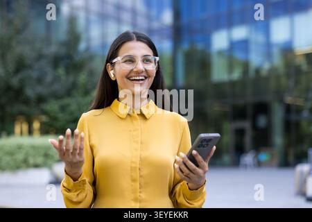 Woman in eyeglasses and yellow shirt smiling happily while interacting on a smartphone. Wearing wireless earphones. Standing outside a modern building Stock Photo