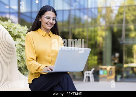 Smiling woman wearing glasses using a laptop while sitting on a modern bench, focusing on her work in a contemporary outdoor office environment Stock Photo