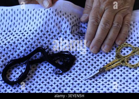 Close-up of hands stitching traditional Japanese Sashiko embroidery in progress on white fabric with black thread and decorative scissors Stock Photo