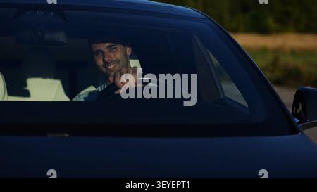Handsome successful entrepreneur looking through windshield of modern electric car. Portrait of young smiling businessman in his innovative electrical Stock Photo