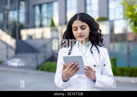 Young indian woman doctor in white lab coat and stethoscope checking patient data on digital tablet, standing outside a modern hospital or clinic building Stock Photo