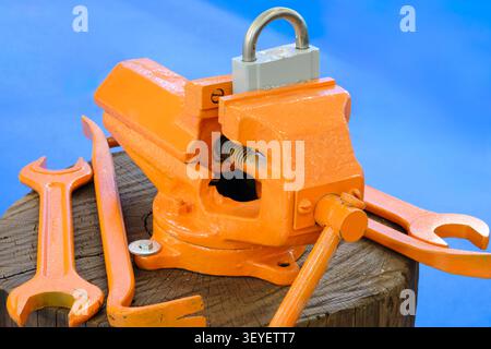 Orange vise secured by a padlock. Wrench and pliers rest on a wooden stump. Blue background contrasts the tools. Industrial equipment close-up view. I Stock Photo