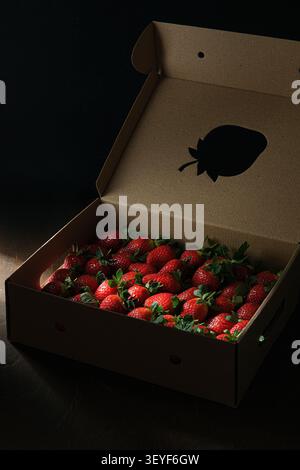 Cardboard box filled with fresh strawberries on dark studio background with dramatic light Stock Photo