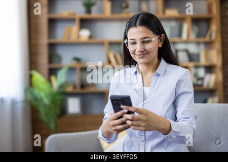 Happy young woman wearing glasses and a blue shirt holding a smartphone, actively sending messages, browsing social media, or checking notifications o Stock Photo