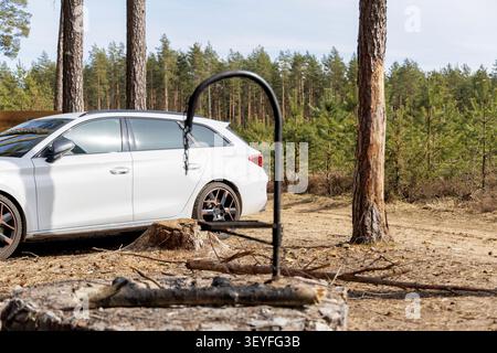 Side angle view of a white estate car parked next to a rustic campfire place and tall pine trees during a sunny outdoor trip. Stock Photo