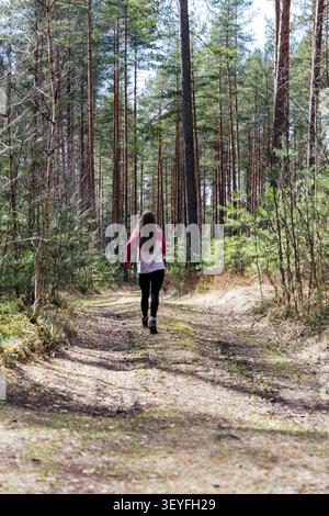 Back view of a young fit woman with long hair running along a sunny dirt path surrounded by tall pine trees. Stock Photo