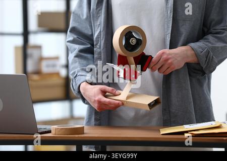 Man using dispenser with adhesive tape to seal cardboard box before sending at table in office, closeup Stock Photo
