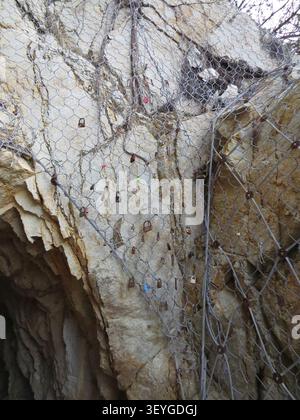 Close view of steep weathered rock wall covered with metal wire mesh and dozens of tiny padlocks, with rough stone texture, cracks, and shadowed reces Stock Photo