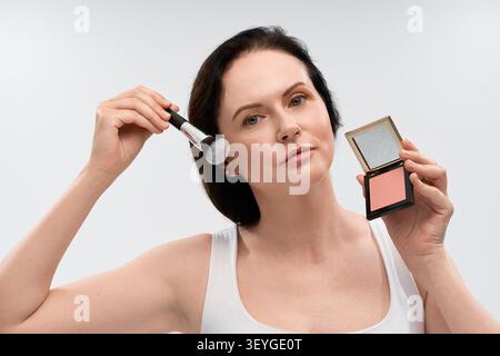 Mature woman showcases her beauty routine, applying blush with grace in a calm atmosphere. Stock Photo