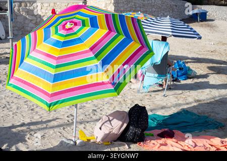 Colorful striped beach umbrella with pink, green, and blue patterns on sandy shore, beach chairs and towels visible in the background Stock Photo