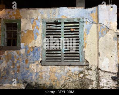 Weathered exterior wall with peeling blue & yellow paint and closed rustic wooden window shutters malaysia Stock Photo