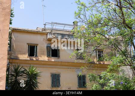 rustic old building facade in southern europe with peeling yellow paint, traditional green window shutters, and lush green trees Stock Photo