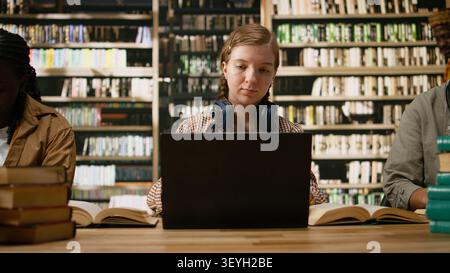 Woman student with headphones typing and working on laptop beside stack of books and blurred shelves in library study space. Digital learning independent study and research. Stock Photo
