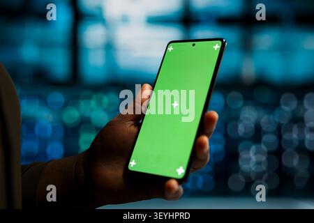 African American system administrator uses a green screen smartphone in a server hub to optimize network resources and reliability. Stock Photo