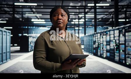 Server facility software engineer checks server facility metrics on a digital tablet to enhance operational effectiveness. Woman in data center working with app device analytics to predict performanc. Stock Photo