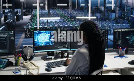 Server facility IT administrator working on AI network software development. African American woman in data center rolling out ML technology working with workstation. Stock Photo