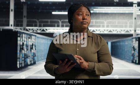 Admin walking in server facility, working with digital tablet to supervise software updates for system stability. Stock Photo