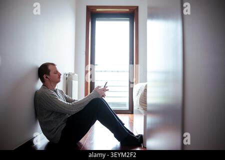 Young man listening to music with headphones and smartphone, leaning on a wall Stock Photo