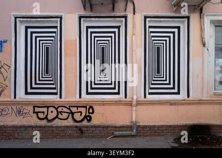 Geometric mural of concentric rectangular patterns painted over three street windows, creating an optical illusion on a worn urban facade Stock Photo