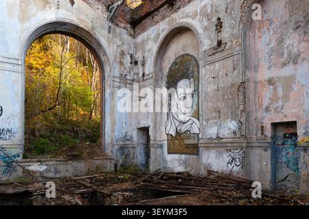 The interior of an abandoned industrial building features a large portrait mural and warm autumn forest light streaming through a tall arched window. Stock Photo