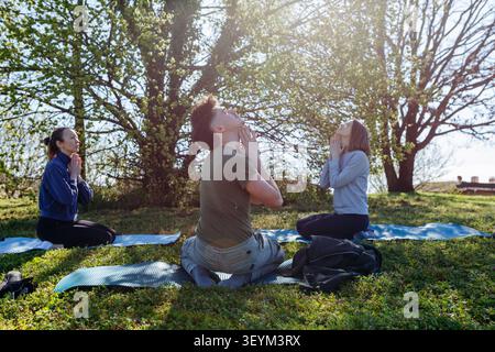 Three adult friends practicing yoga and mindfulness together outdoors in a sunny park Stock Photo