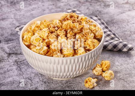 Close-up of golden caramel popcorn in textured white bowl, scattered kernels on gray stone table, checkered napkin background, snack food photography Stock Photo