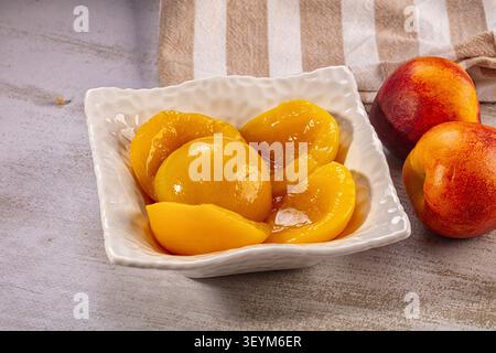 Glossy yellow peach slices in textured white bowl beside two whole ripe peaches on stone surface with striped cloth background Stock Photo
