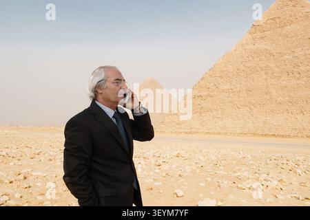 Focused Arab executive in dark suit making a decisive cell phone call at the iconic Great Pyramids symbolizing global leadership authority Stock Photo