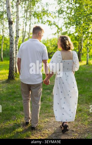 A young couple walks hand-in-hand on a dirt path surrounded by birch trees and green grass, bathed in warm sunlight Stock Photo
