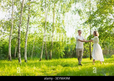 A happy young couple holding hands and twirling in a vibrant green birch grove on a bright sunny day, surrounded by lush foliage and dappled light Stock Photo