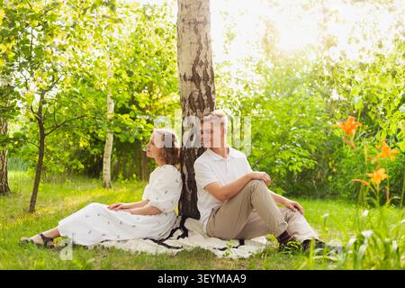 A young man and woman enjoy a peaceful moment together, leaning against a tree trunk on a blanket amidst lush greenery and bright sunlight Stock Photo