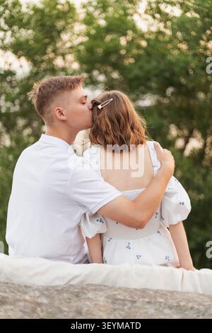 A young man gently kisses the forehead of his partner while embracing her from behind, surrounded by nature's soft glow Stock Photo