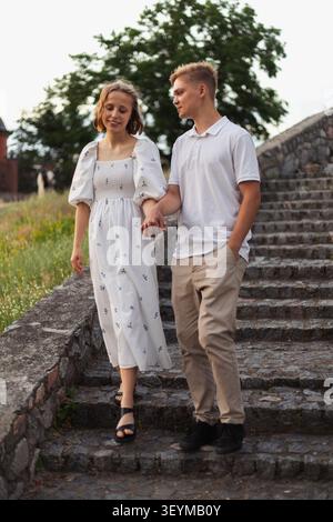 A teenage couple walks hand-in-hand down a worn stone staircase surrounded by greenery and an old stone wall Stock Photo
